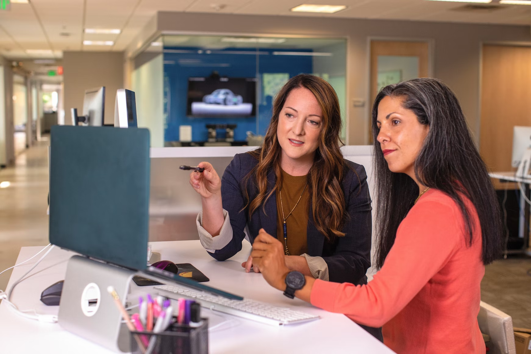Two women are seated at a desk looking at a laptop screen. One woman is pointing at an object on the screen while the other looks on. 